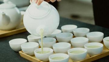 Pouring tea into multiple small white cups arranged on a wooden tray.