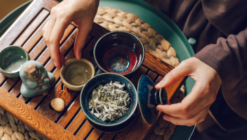 Hands arranging tea set and herbs on a wooden tray with a small statue.