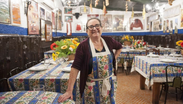 Smiling woman in apron stands in decorated restaurant with colorful tablecloths and flower arrangements.