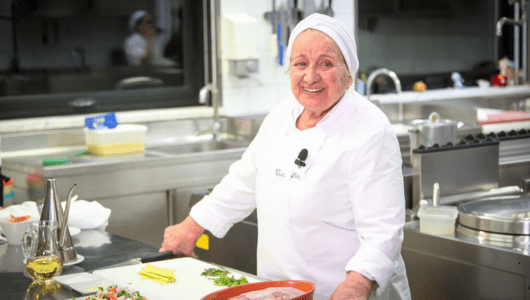 Elderly chef with white uniform smiling in a professional kitchen.