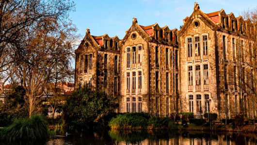 Vintage stone building with gabled roofs by a pond and surrounded by bare trees.