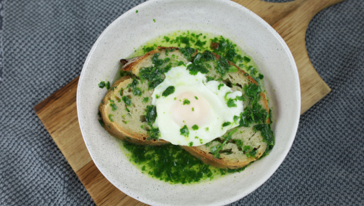 Poached egg on sliced bread with green sauce in a white bowl on a wooden board.