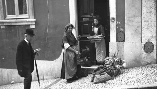 Two women exchange goods at a doorway; a man walks past with a walking stick.