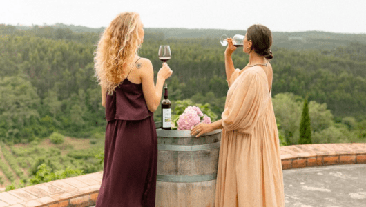 Two women with wine glasses stand by a barrel overlooking a scenic forest.