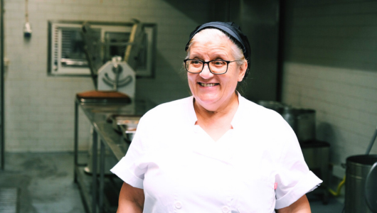 Smiling woman in a kitchen wearing a white chef uniform and black hair net.