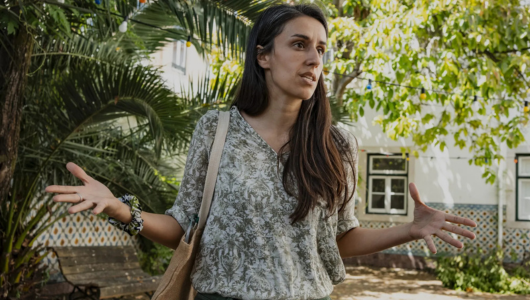 Woman with long hair gestures while speaking outdoors near trees and a building.