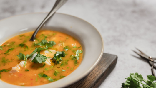 Bowl of orange soup with cilantro garnish and a spoon on a wooden board.