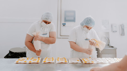 Two workers in protective clothing piping food onto sheets in a clean workspace.