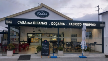 Small café with 'Casa das Bifanas' sign and outdoor seating, umbrellas folded.