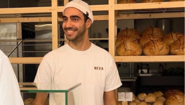 Smiling man in bakery with wooden shelves and bread loaves in background.