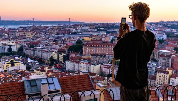 Person photographing a cityscape at sunset with a bridge and rooftops in view.