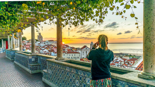 Person photographing a scenic sunset view over tiled terrace roofs and ocean.