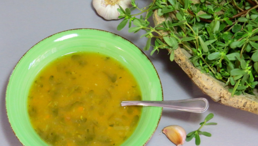 Green bowl of orange soup with herbs next to garlic and a basket of greens.