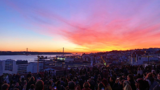 Crowds gather overlooking a cityscape at sunset with a vibrant sky.