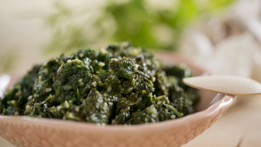 Close-up of a bowl with green herb mixture, wooden spoon beside.