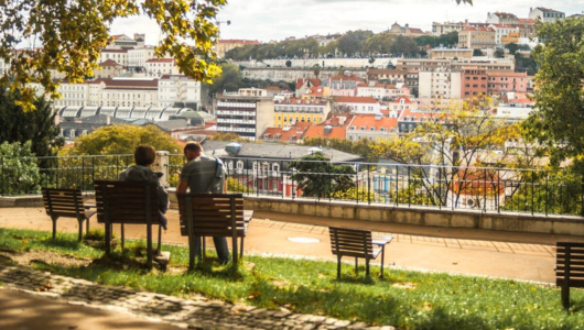 Two people sit on benches overlooking a cityscape with trees and buildings.