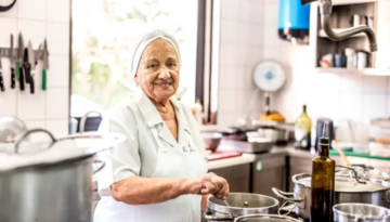 Smiling elderly woman cooking in a kitchen with pots and a bottle on the counter.
