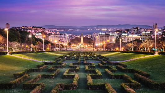 A sunset view of an illuminated park with a geometric hedge design and cityscape in the background.