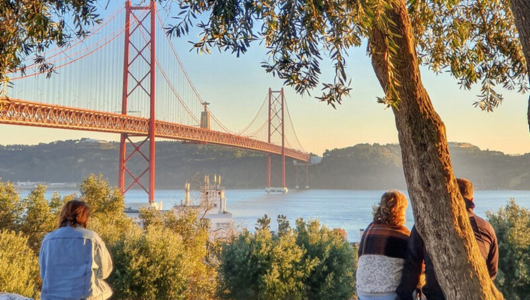 Three people sitting near trees, overlooking a large red suspension bridge over a river.