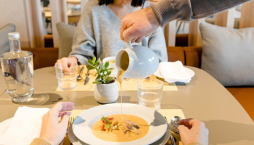 Person pours soup into a bowl on a dining table with two seated individuals.