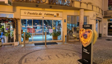 Night view of a pastry shop with 'O Pasteís de Fátima' sign and heart-shaped pastry display outside.