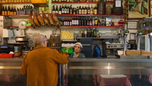 Man at bar counter with shelves of bottles and hanging meats in a cozy deli or café.