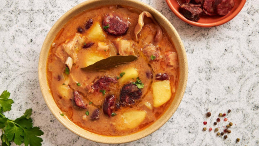 Bowl of stew with potatoes, sausage, and herbs on a textured surface.