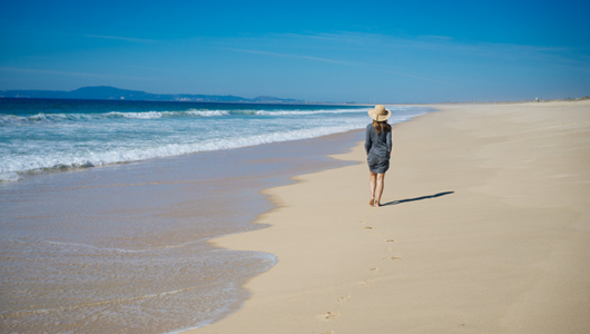 Person in hat walking alone on a sandy beach under a clear blue sky.