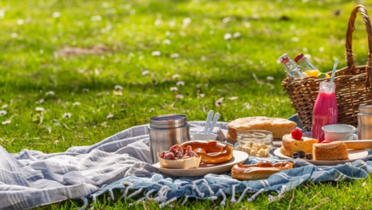 Picnic scene with food on a blanket, basket, and drinks on a sunny lawn.
