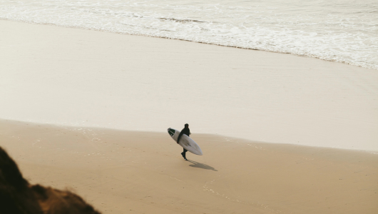 Lone surfer walking with surfboard on a sandy beach towards the ocean.