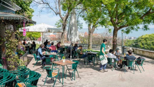 People seated at outdoor cafe with green chairs and trees.