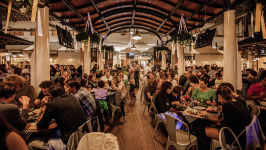 Crowded indoor restaurant with people dining at long tables.