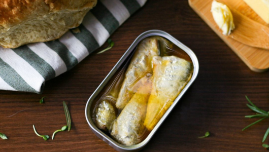 Open can of sardines on wooden table with bread and butter.
