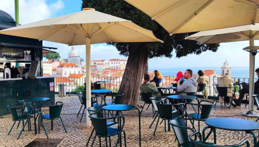 Outdoor cafe with tables, chairs, and umbrellas, overlooking a cityscape and ocean view.