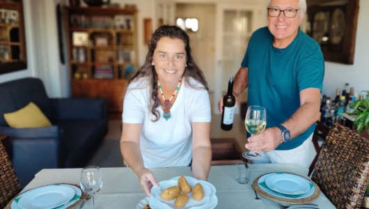 Two people setting a dining table with wine and food in a cozy room.