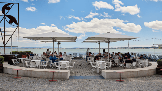 Outdoor cafe by the sea with people seated under large umbrellas.