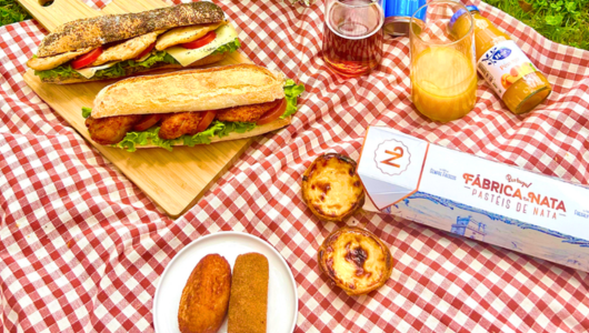 Picnic scene with baguette sandwiches, pastries, juice, and sauce on a red checkered cloth.