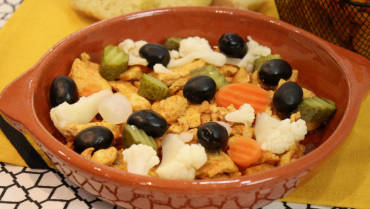 Ceramic dish with chicken, olives, pickles, cauliflower, and carrots on yellow and black cloth.