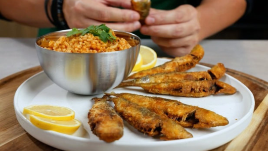 Person holding food near plate with fried fish, bulgur in a bowl, and lemon slices.