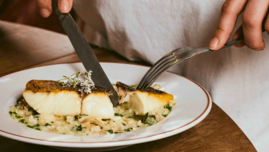 Person cutting fish on risotto on white plate with fork and knife.