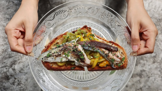 Hands holding a plate with fish-topped toast and vegetables.