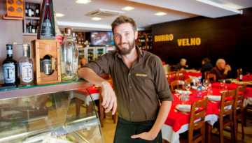 Man in a restaurant with red tablecloths, standing near a display case with bottles.