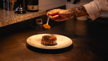 Chef's tattooed hand drizzling sauce on meat dish on a white plate.