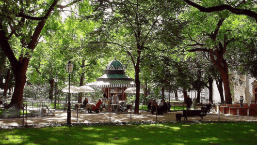 People sitting at tables under trees in a park with a gazebo.