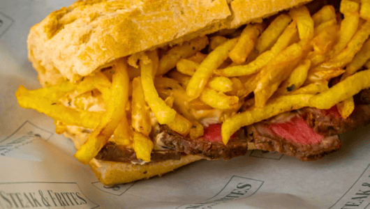 Sandwich filled with steak slices and French fries on paper.