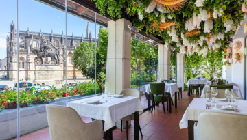 Elegant restaurant with hanging greenery, white tablecloths, and large windows overlooking a historic building.