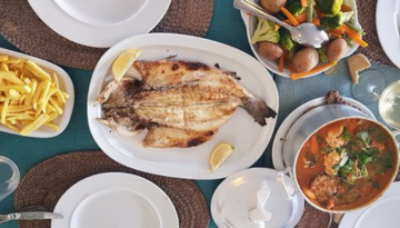 Table with grilled fish, fries, salad, and potatoes in bowls.