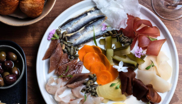 A charcuterie platter with fish, vegetables, seeds, and flowers on a white plate.