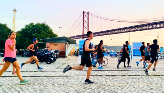 People jogging on a waterfront path near a large bridge at sunset.