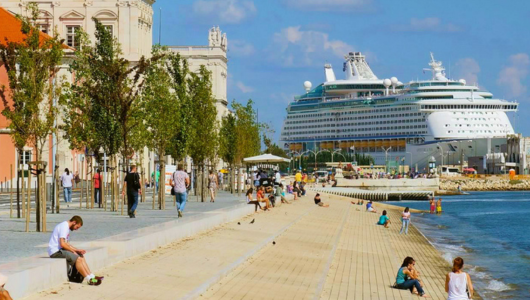 People walking along a waterfront promenade with a large cruise ship docked nearby.